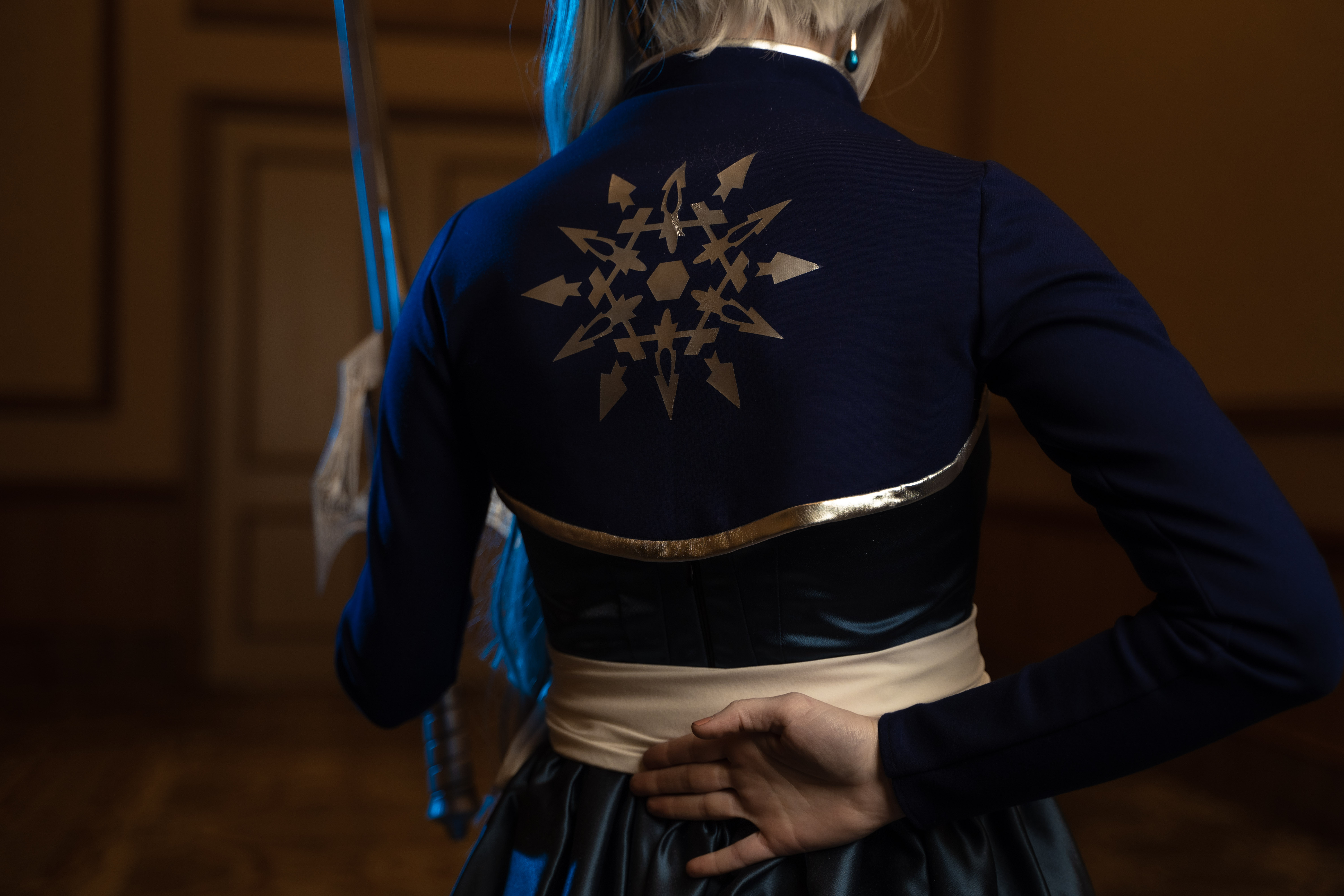 Female character with silver/white ponytail and tiara, dressed in a blue ombre dress and bolero with large silver snowflake on back. Waist is wrapped in white fabric belt and she carries a silver sword.