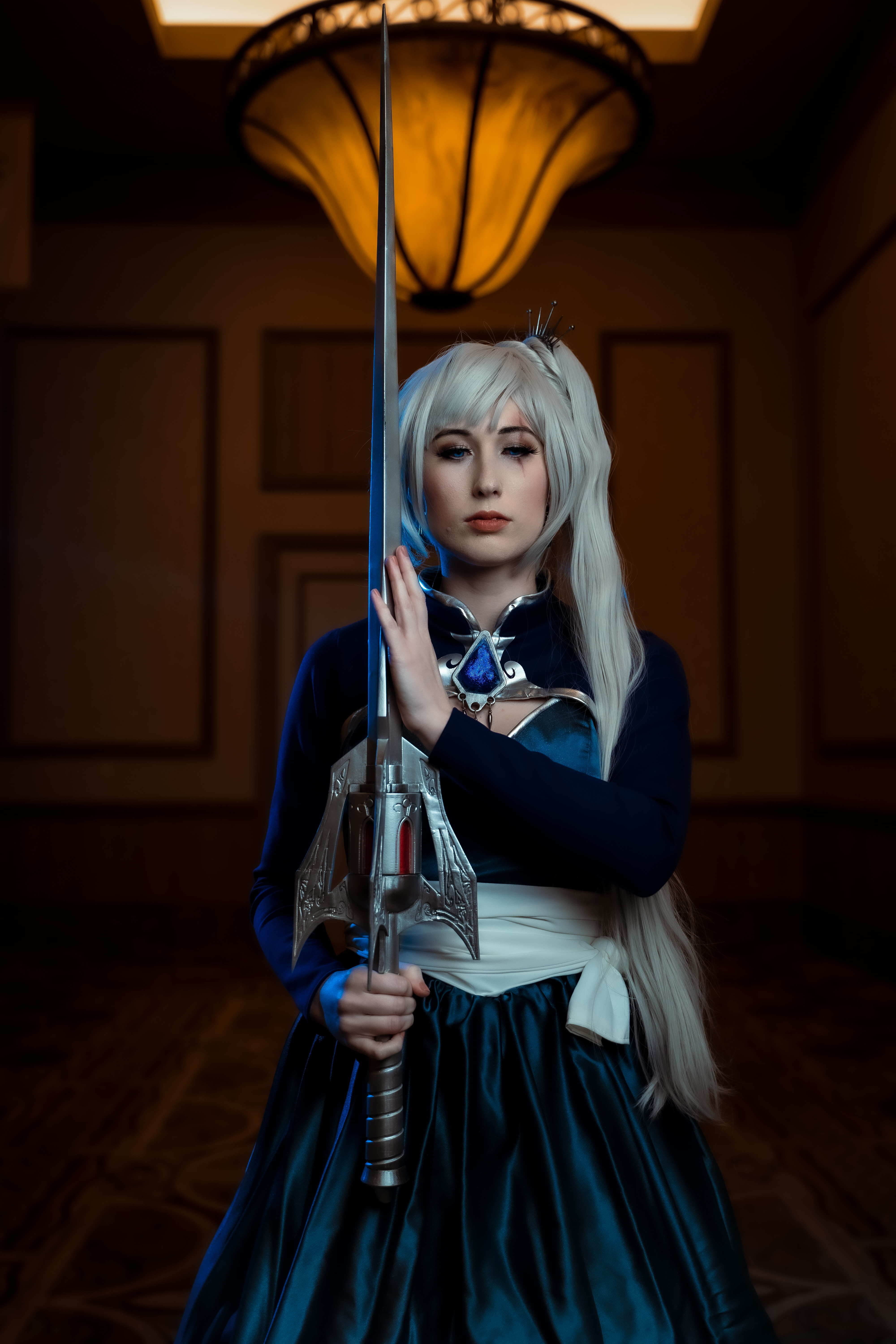 Female character with silver/white ponytail and tiara, dressed in a blue ombre dress and bolero with large silver snowflake on back. Waist is wrapped in white fabric belt and she carries a silver sword.