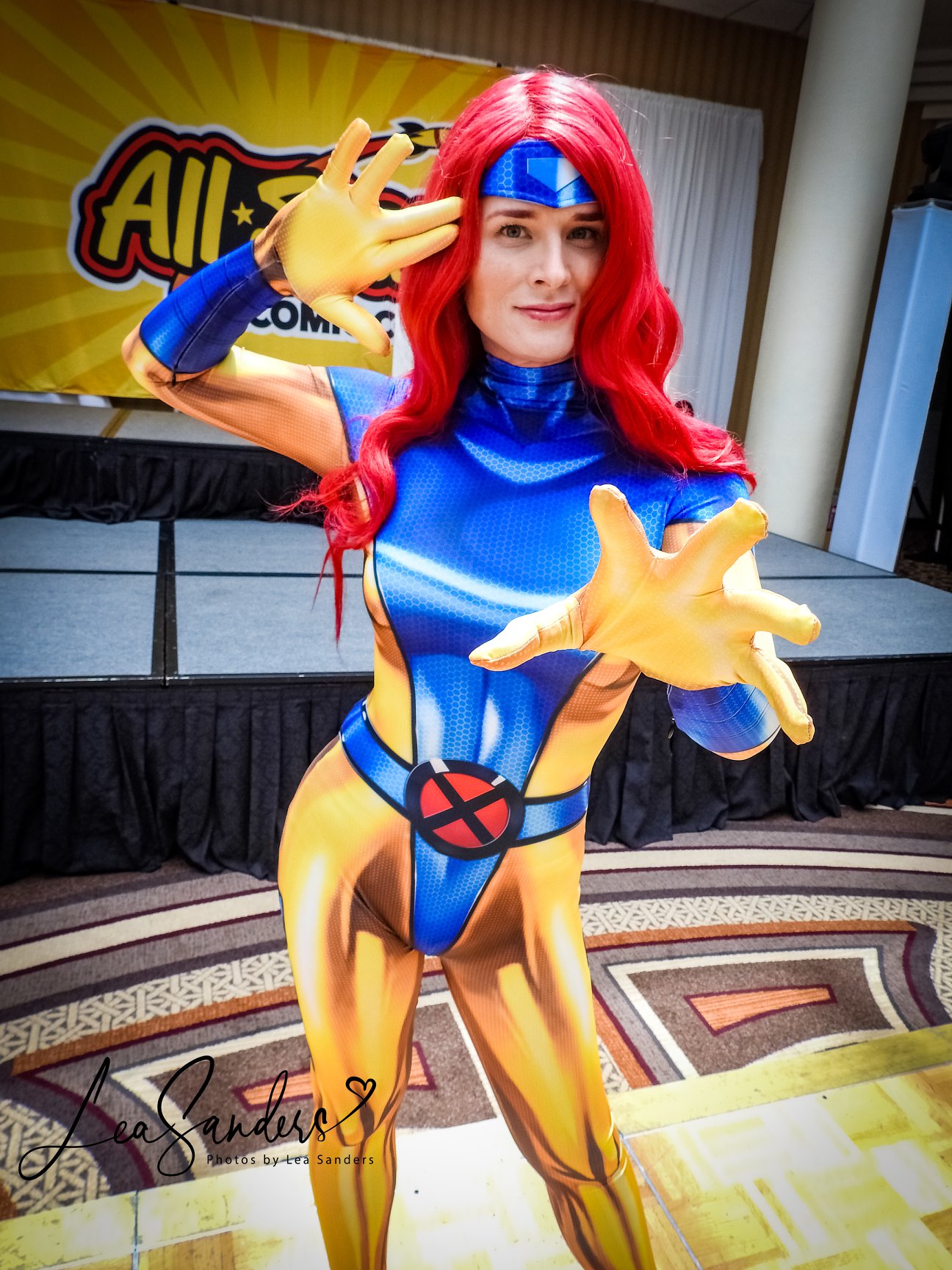 Jay is wearing a cosplay of Jean Grey's 1990s blue and yellow X-men uniform from the comic and animated series. She has one hand to her temple and one hand reaching toward the camera, as if reading your mind.