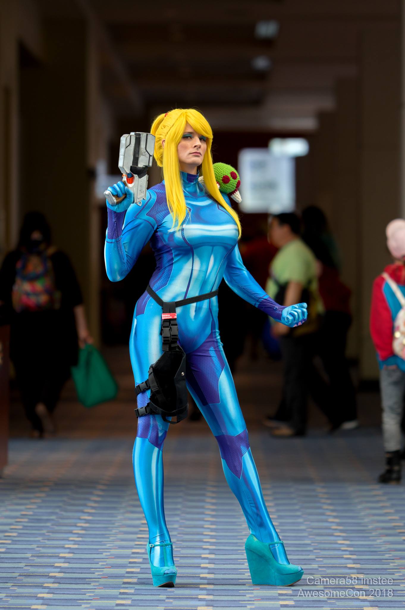 Jay is wearing a cosplay of Samus Aran's blue Zero Suit from the Metroid game series. She is holding a paralyzer pistol in one hand and making a fist with the other, while a baby metroid rests on her shoulder.
