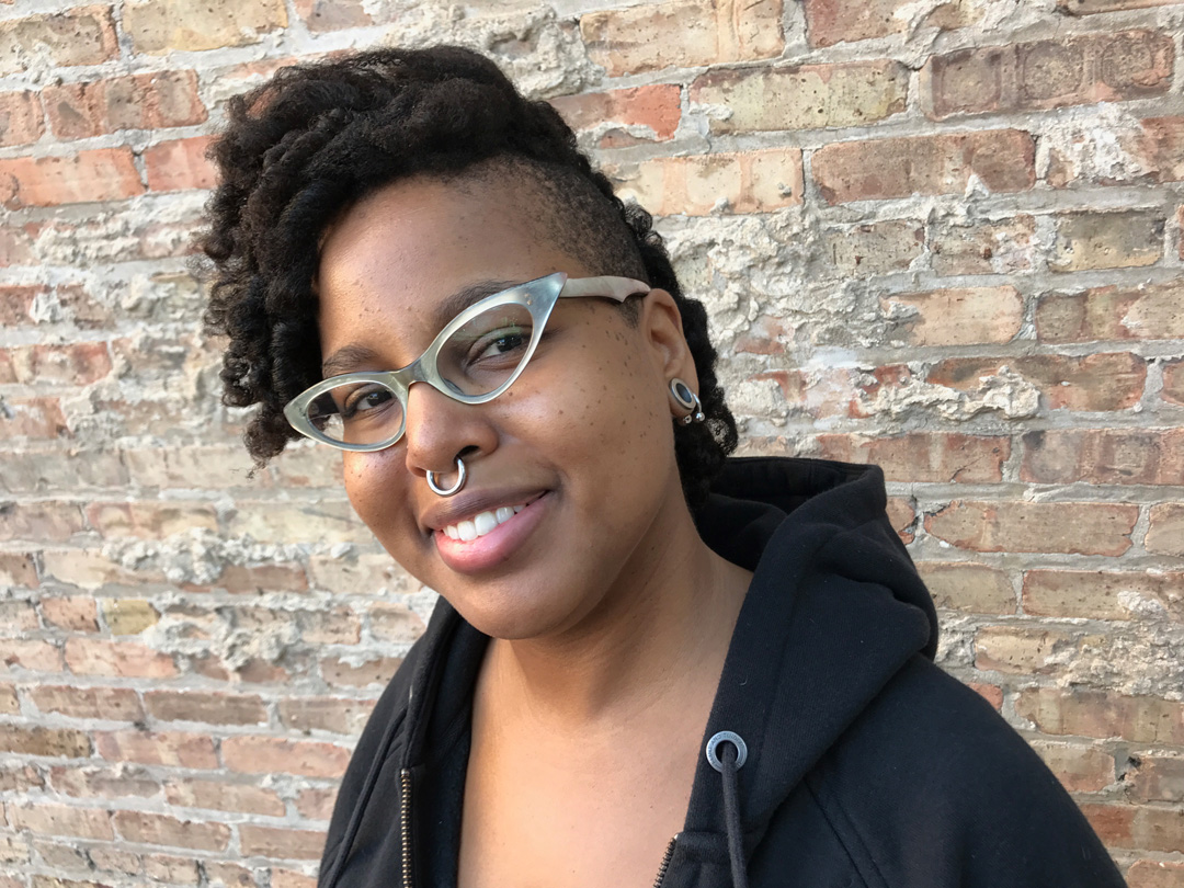 Photo of C. Spike Trotman, artist, writer, and publisher, a black woman with a nose ring and light-colored classes and her hair partially shaved on the side. She is smiling towards the camera.