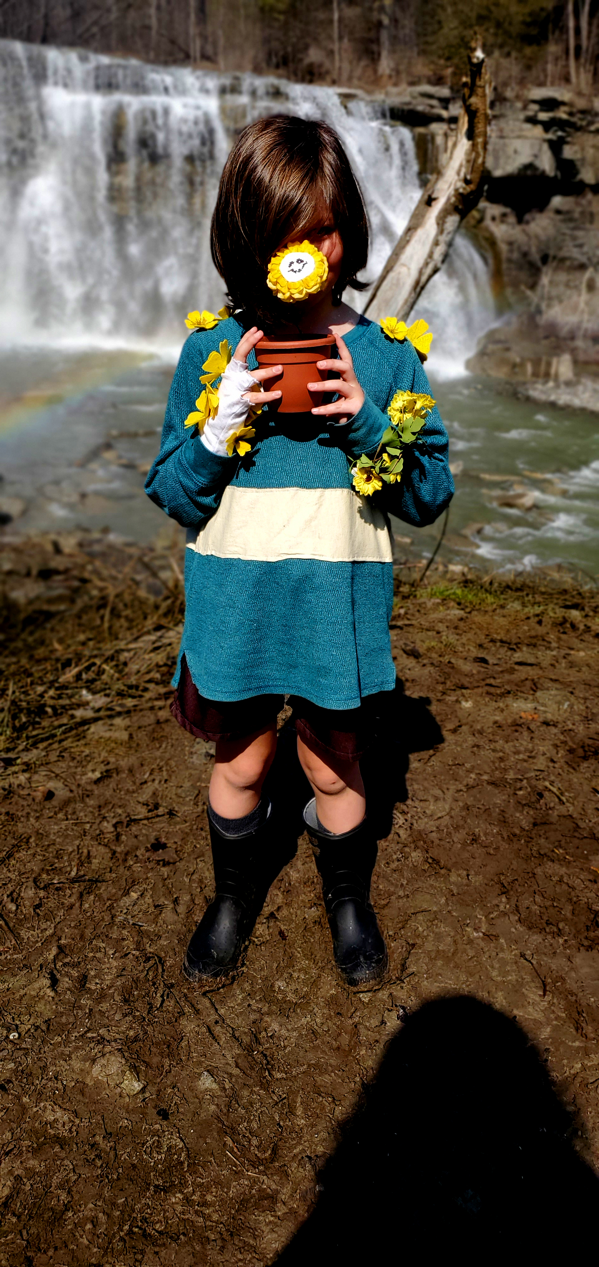 A small white child with brown hair, a green shirt with one yellow stripe, brown shorts and brown boots with an ominous smile.