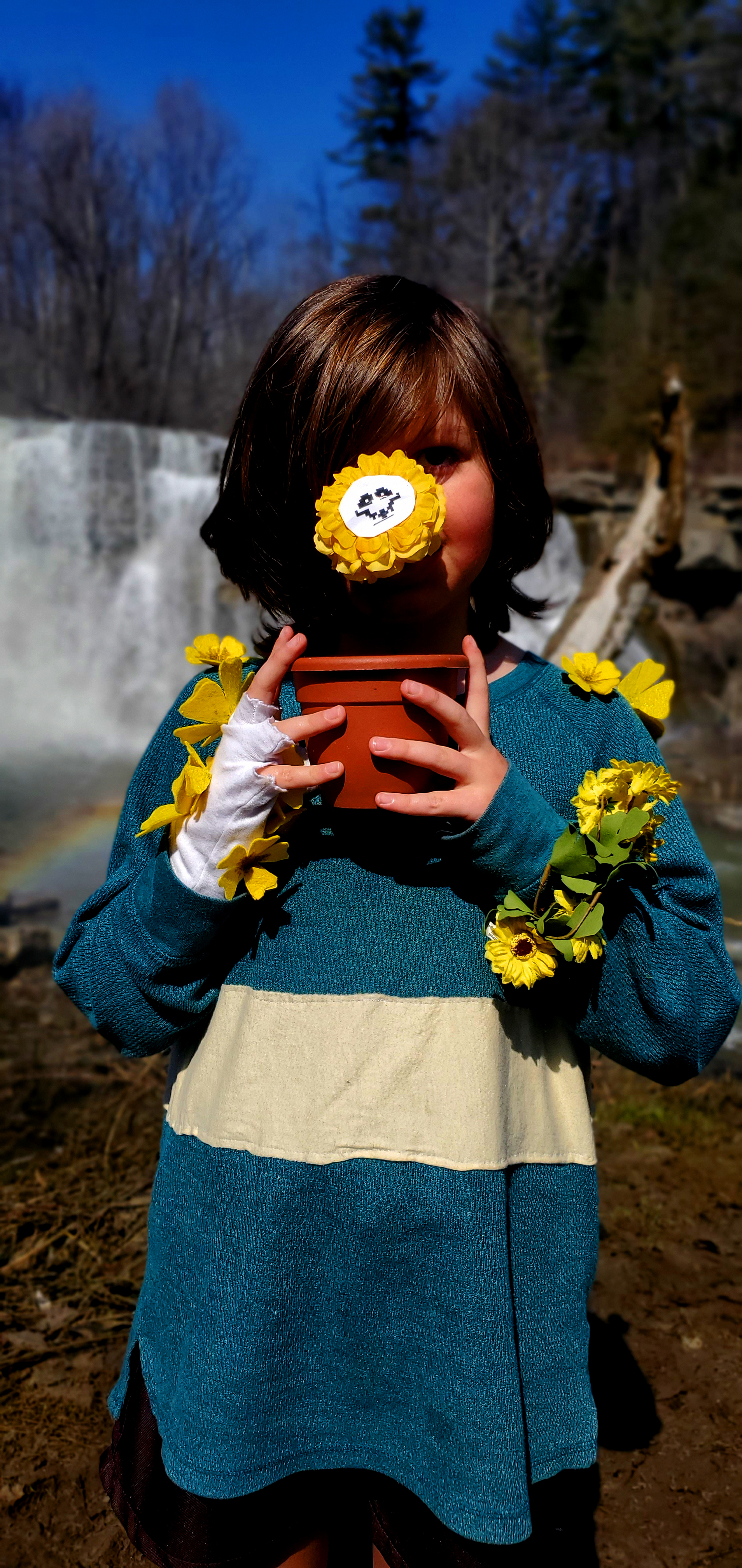 A small white child with brown hair, a green shirt with one yellow stripe, brown shorts and brown boots with an ominous smile.