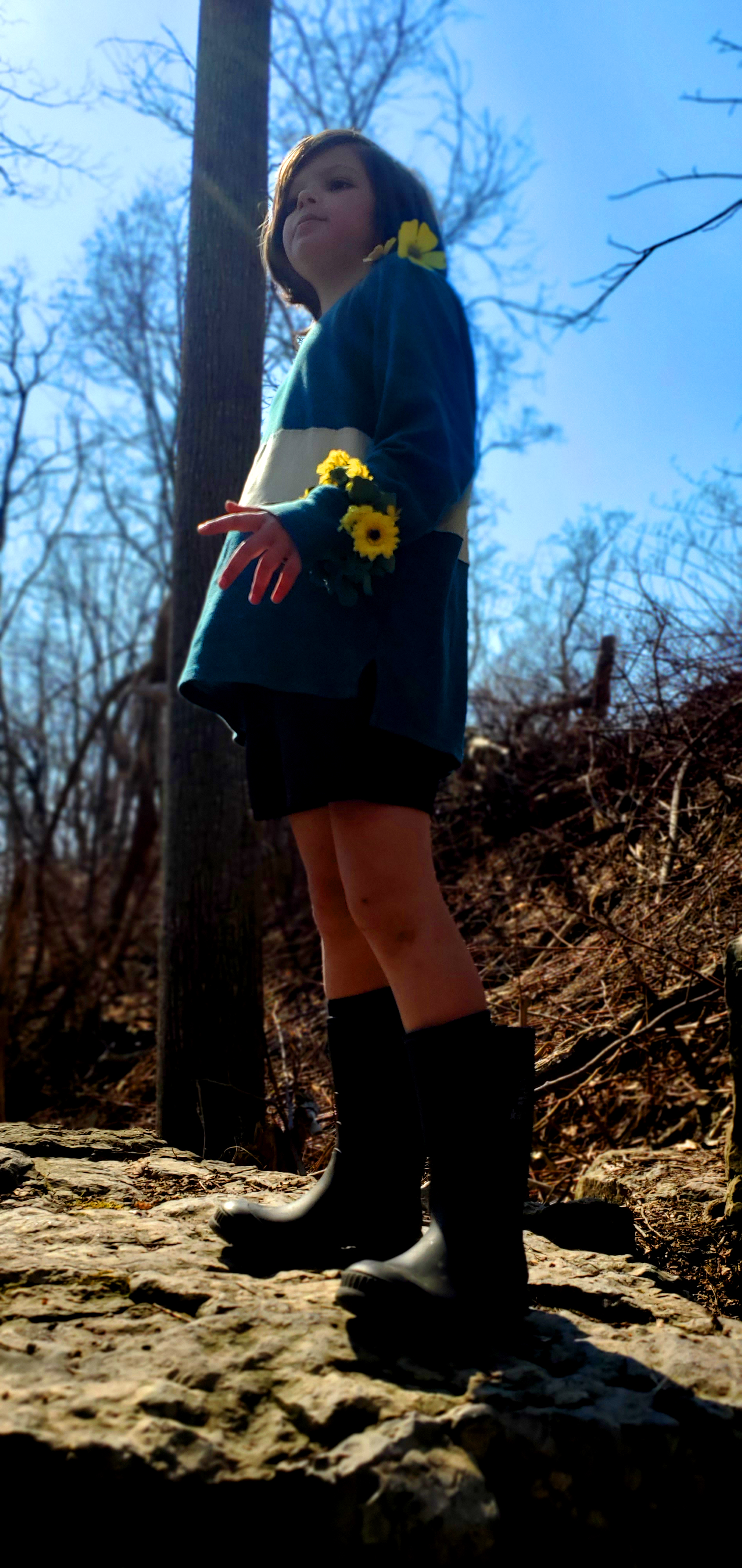 A small white child with brown hair, a green shirt with one yellow stripe, brown shorts and brown boots with an ominous smile.