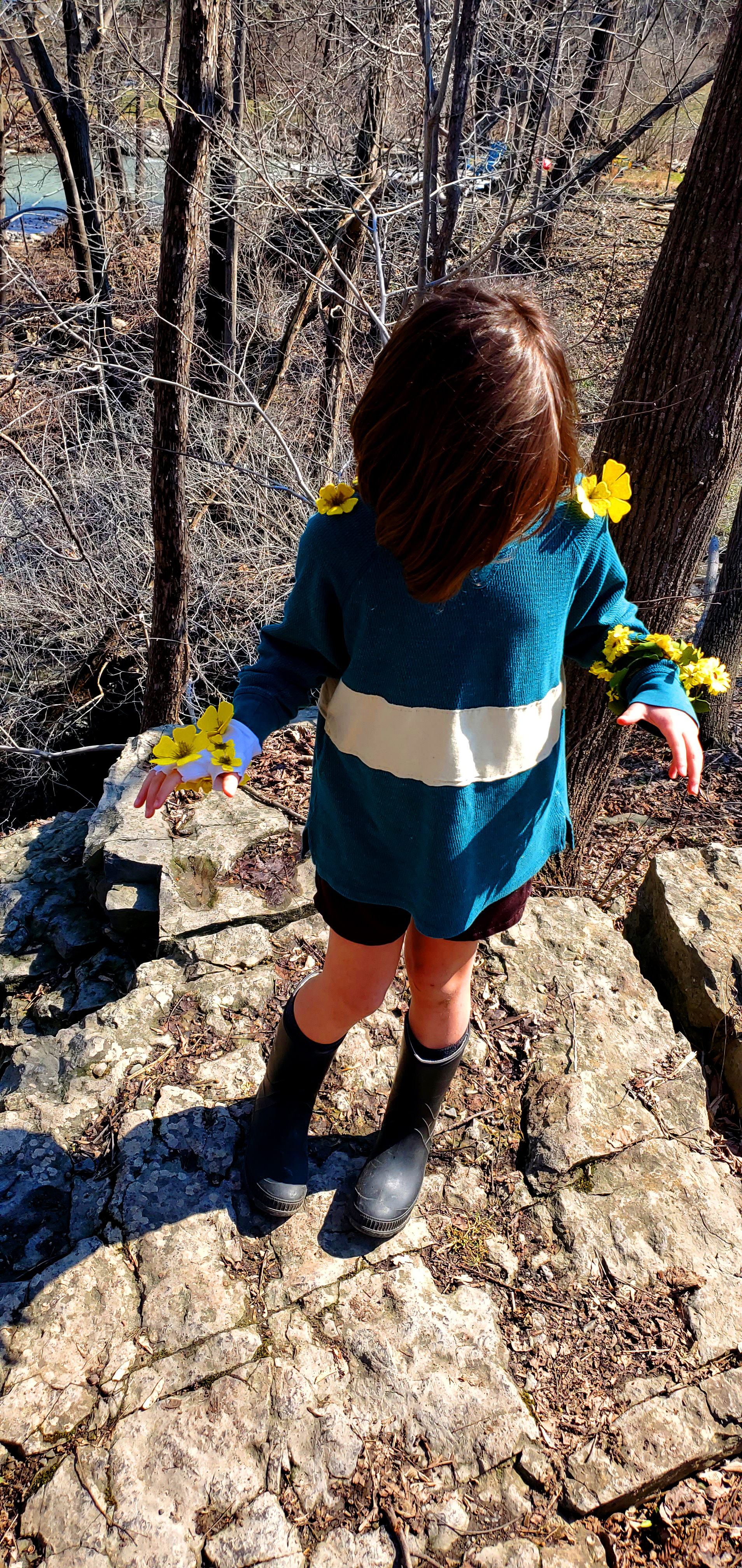 A small white child with brown hair, a green shirt with one yellow stripe, brown shorts and brown boots with an ominous smile.