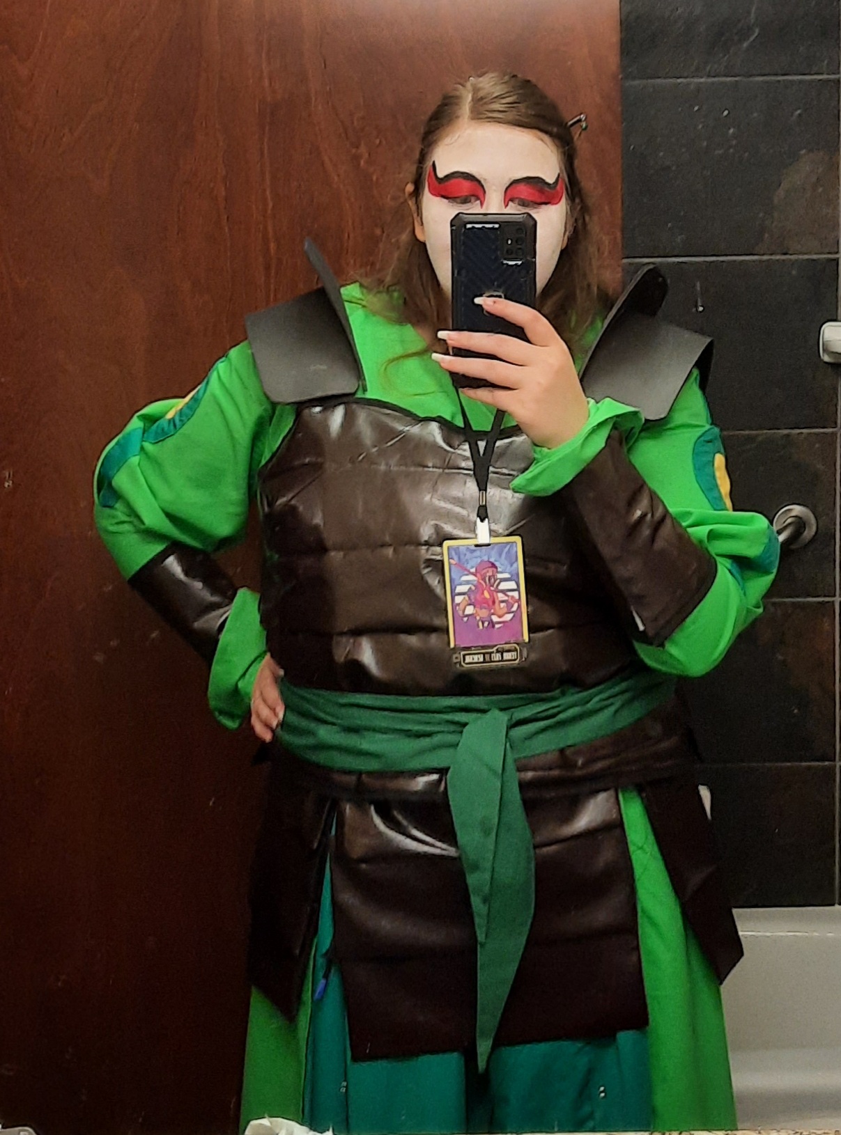 Three female cosplayers. They are dressed in dark green skirt with diagonal pleats, light green long jackets with long poofy sleeves, and leather (plether) armor and wrist guards. They have white face paint with red eye makeup. Due to covid had to wear white masks with red lips to stay safe but keep the look. They are holding large wooden fans.