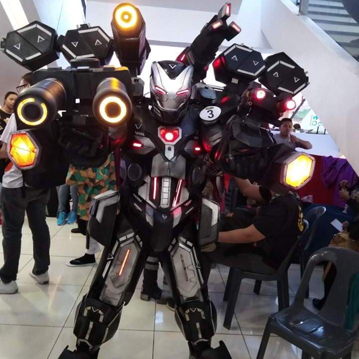 War Machine, in grey armor, stands with both arms full of guns and lights.