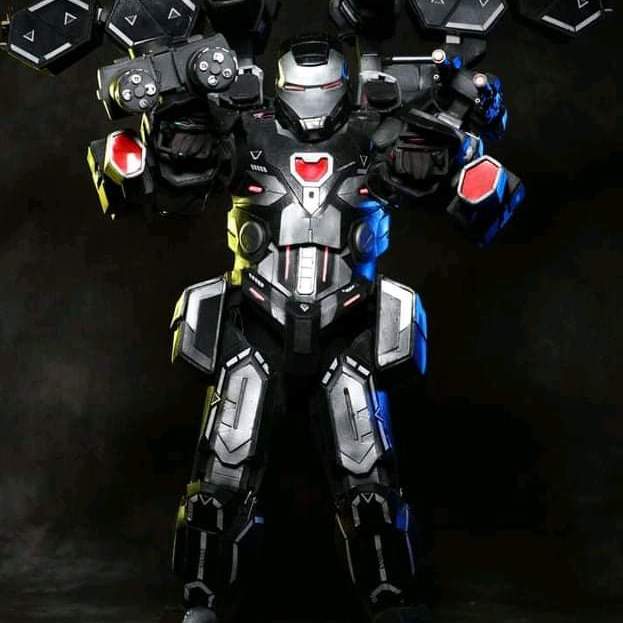 War Machine, in grey armor, stands in a black space with lit up guns.
