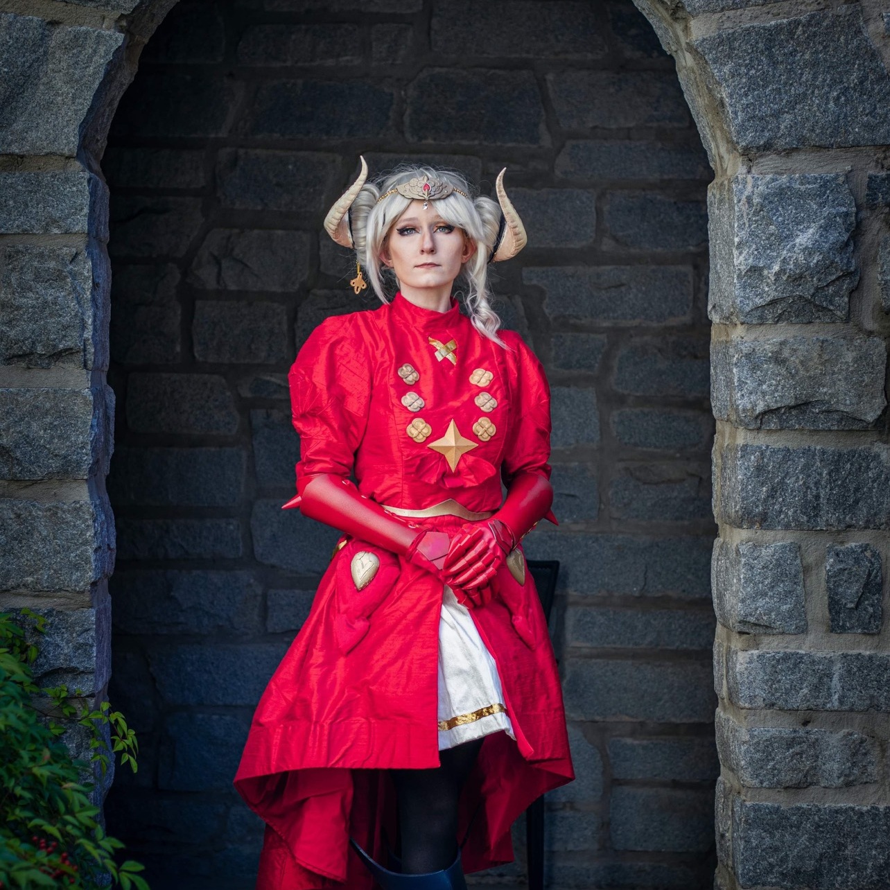 Woman with horns in a red cape and tunic in front of an old building.