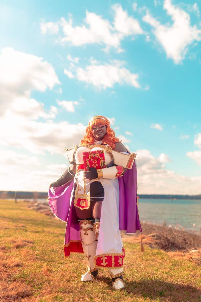 Black and plus sized woman with eyes closed wearing orange wig with white and gold headband, wearing a white over dress with red and gold details and purple lining, white cape with details and purple lining, white chest, hanging shoulder and bracer armor with red and gold details, and knee, bracers, shoe, & grieves armor with gold details. She's also wearing a black short skirt underneath, black thigh highs, and black fingerless gloves. Holding a red book with black and gold details on the left arm while holding the tip of the cape on her right hand. Standing on grass with the ocean and sky on the background.