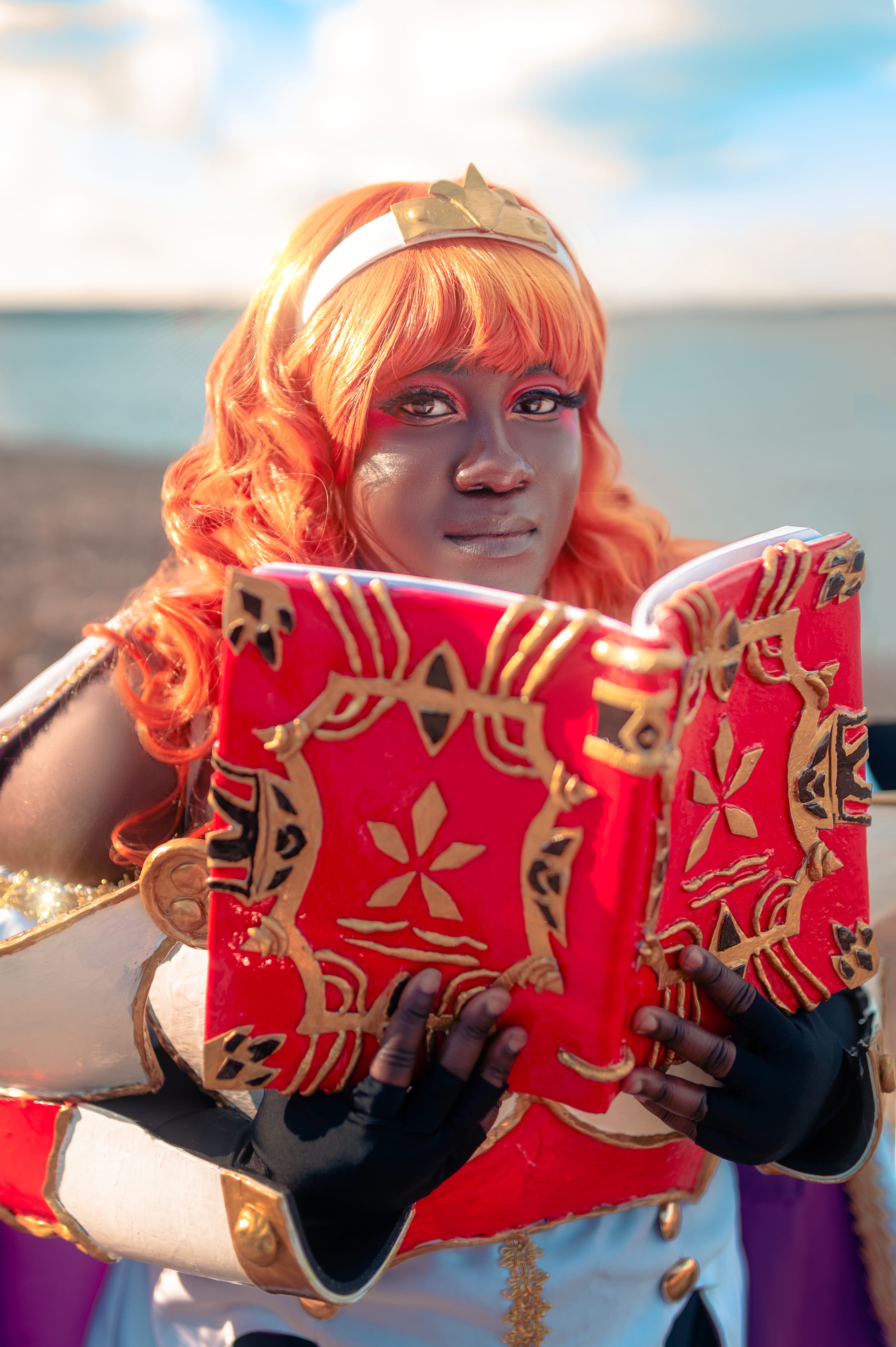 Close up of black and plus sized woman from the waist up wearing orange wig with a white and fold headband. She's also wearing the white dress with gold details and white cape with gold details. she's also wearing white chest and bracers armor with red and gold details. She's also wearing white hanging shoulder armor and black fingerless gloves.. She's holding a red book with gold and black details opens it showing both sides of the book while facing the camera.
