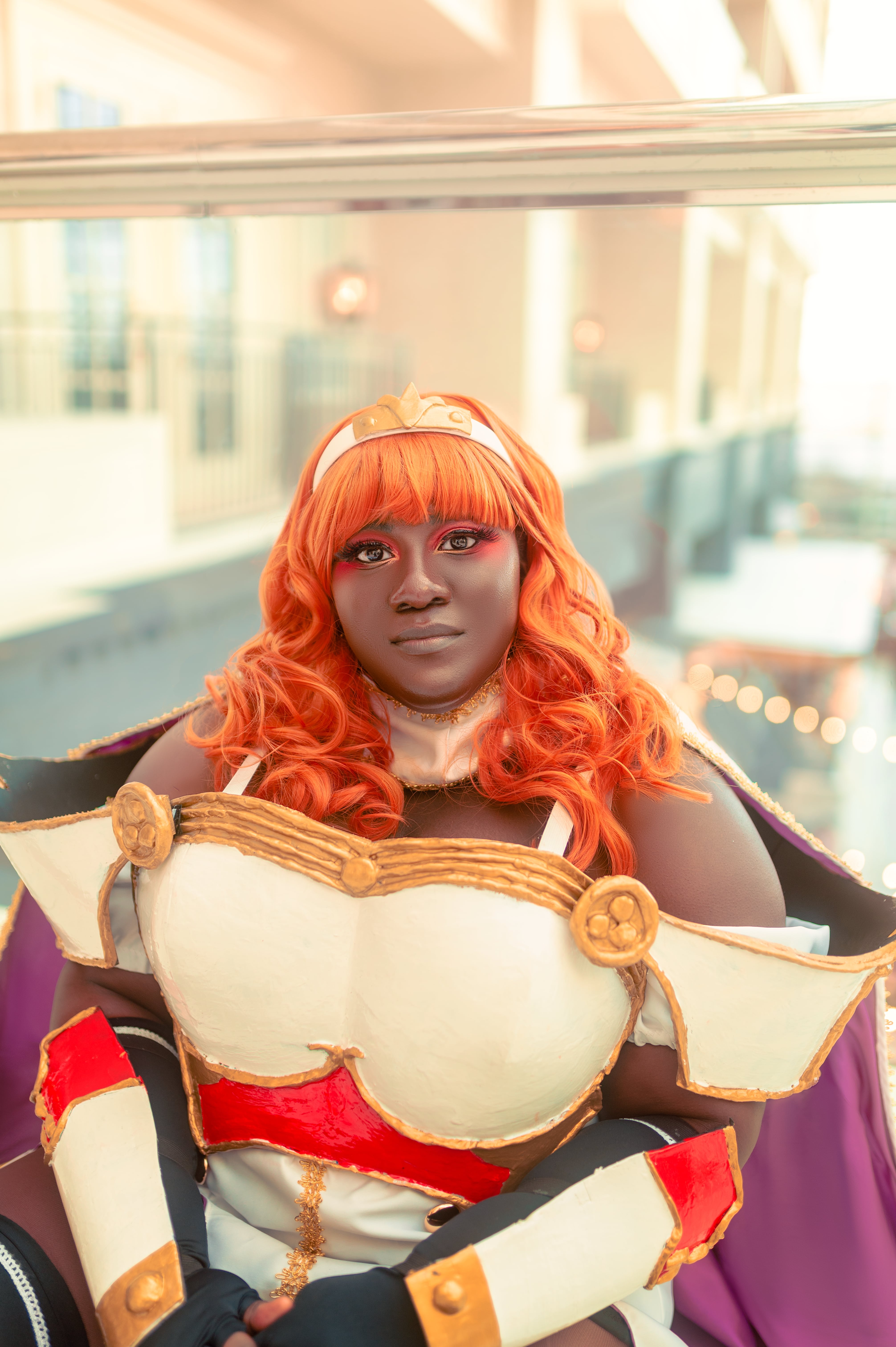 Black and plus sized woman from the waist up facing the camera. She's wearing an orange wig with white and gold headband. She's also wearing a white dress and cape with gold details, white chest, hanging shoulder, and bracer armor with red and gold details. She's sitting down with her hands together on her lap and facing the camera.
